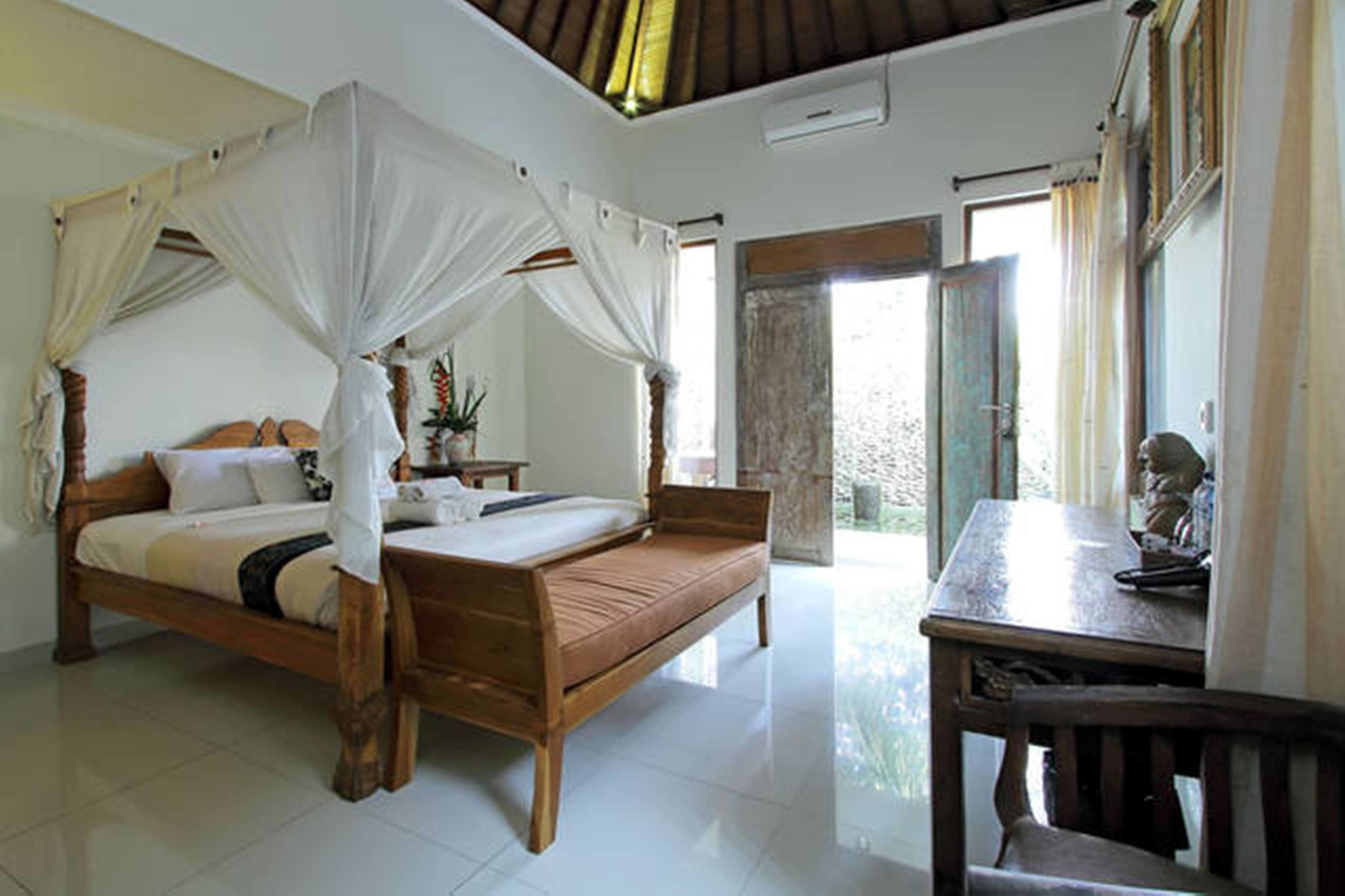 A room with a wooden bed, a small table, and a chair. There is a window with curtains and a view outside.