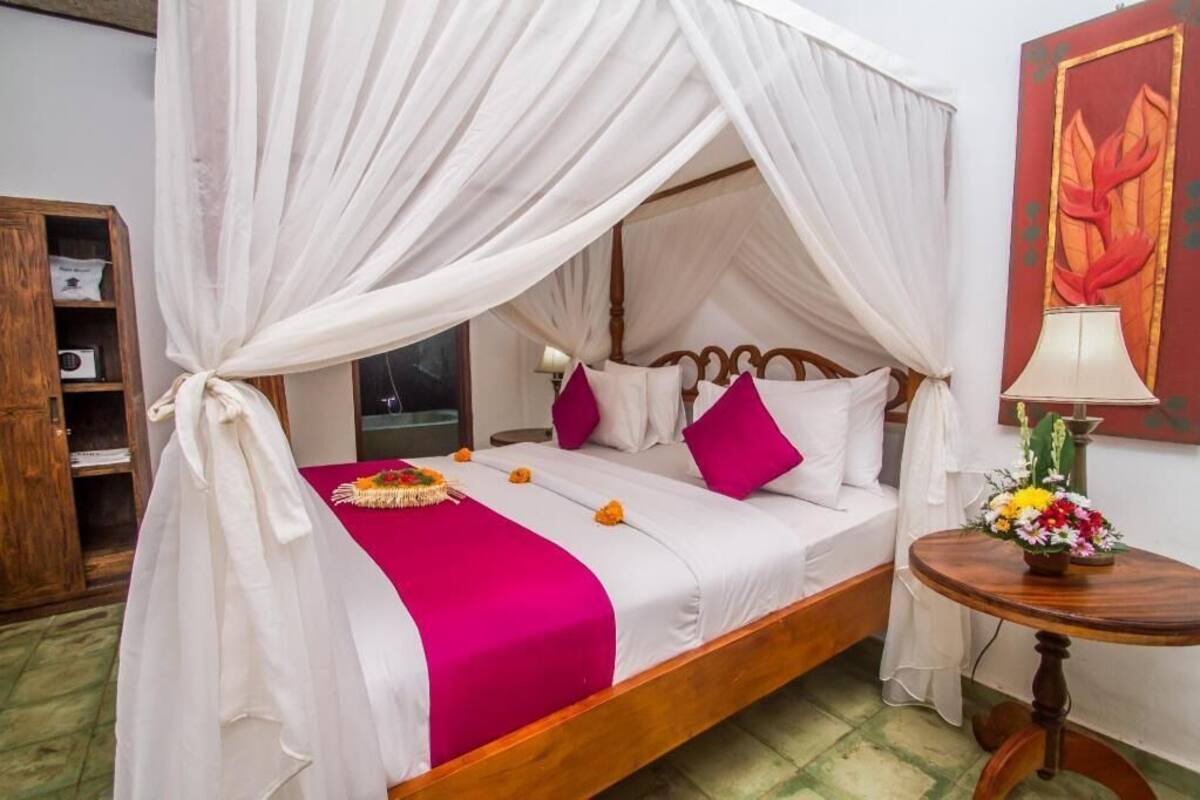 A four-poster canopy bed with white bedding and a red throw blanket, a wooden nightstand with a lamp, and a wall-mounted painting.
