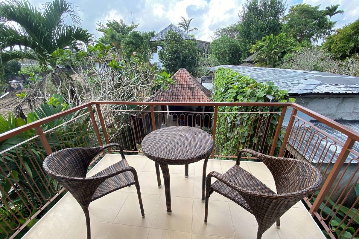 A balcony with wicker furniture and a tiled floor, surrounded by greenery and a building in the background.