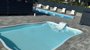 Une piscine à bord incurvé, entourée de carreaux gris. On y voit des chaises longues blanches et, au fond, un palmier.