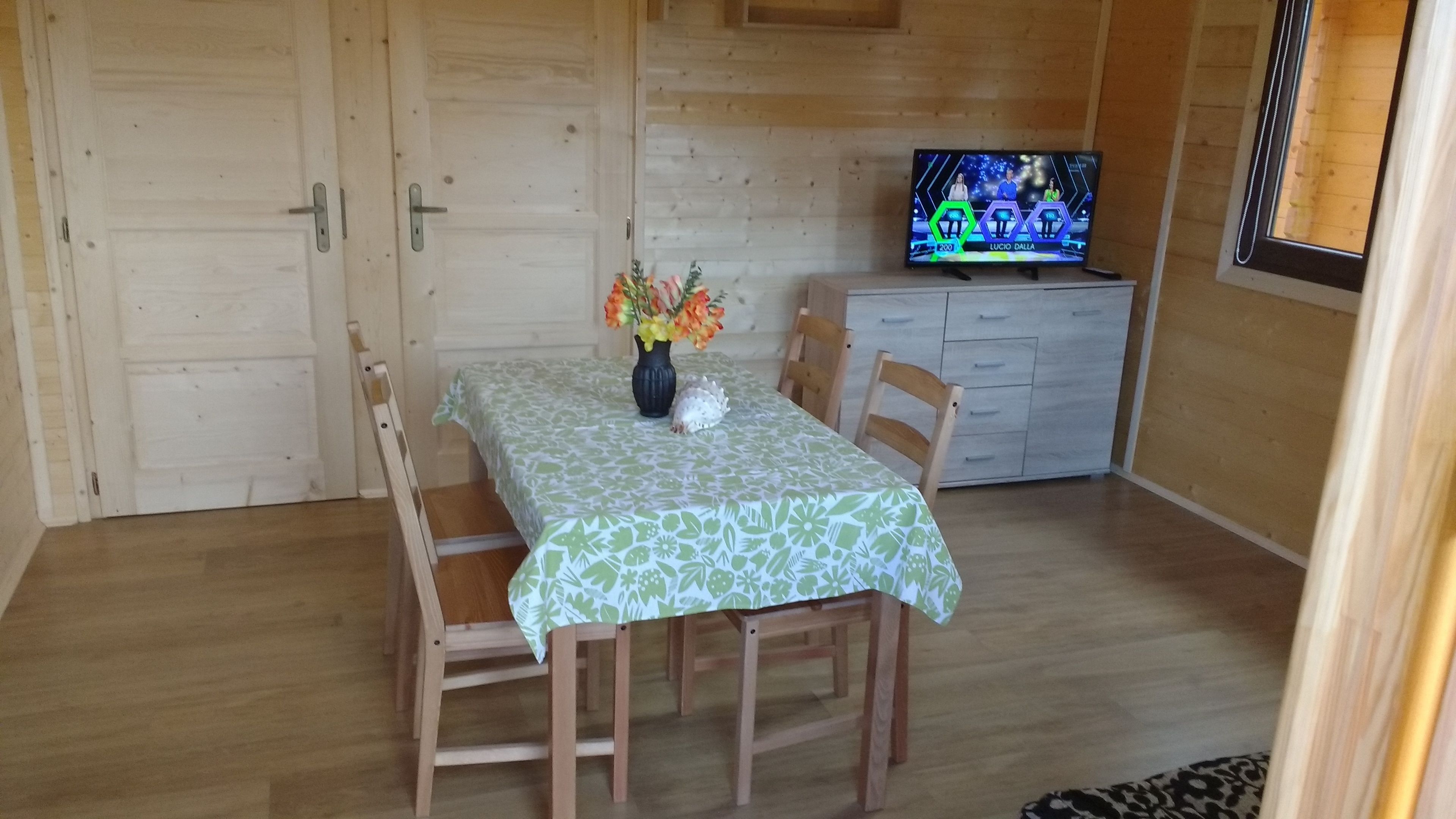Ein Holzinterieur mit einem Esstisch, der mit einer grün-blumigen Tischdecke bedeckt ist, zwei Holzsesseln, einer Blumenvase, einem Fernseher auf einem Schrank und einem Fenster mit Jalousien.