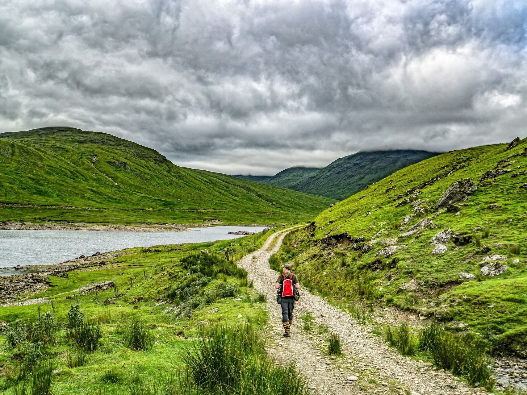 A person walking on a gravel path surrounded by green hills and a lake.