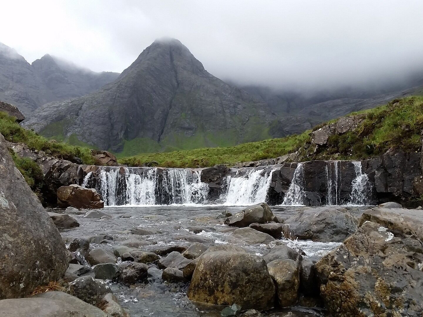 A waterfall cascading over rocks in a mountainous area with a prominent peak in the background.