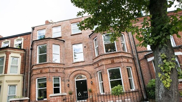A multi-story brick building with a black front door, a small garden, and a tree in the foreground.