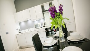 A modern kitchen with white cabinets, a black dining table set with white plates, bowls, and cutlery, and a vase with purple flowers.