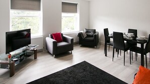 A modern living room with a flat-screen TV, two sofas, a dining table with chairs, and large windows with blinds.
