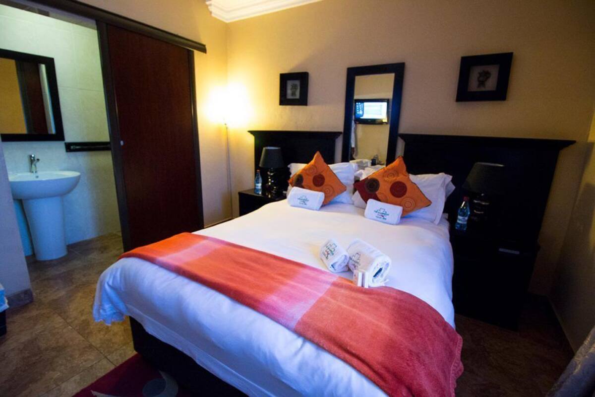 A hotel room with a bed, two pillows, a nightstand, a lamp, a bedside table, a mirror, and a bathroom with a sink and toilet.