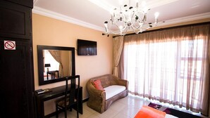 A hotel room with a bed, a desk with a chair, a large mirror, a television on the wall, and a window with curtains. - The Shamrock Lodge (Polokwane)