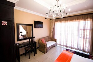 A hotel room with a bed, a desk with a chair, a large mirror, a television on the wall, and a window with curtains. - The Shamrock Lodge (Polokwane)