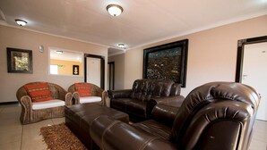 A living room with a brown leather sofa, a wicker armchair, and a matching ottoman. There are framed pictures on the walls and a large window in the background. - The Shamrock Lodge (Polokwane)