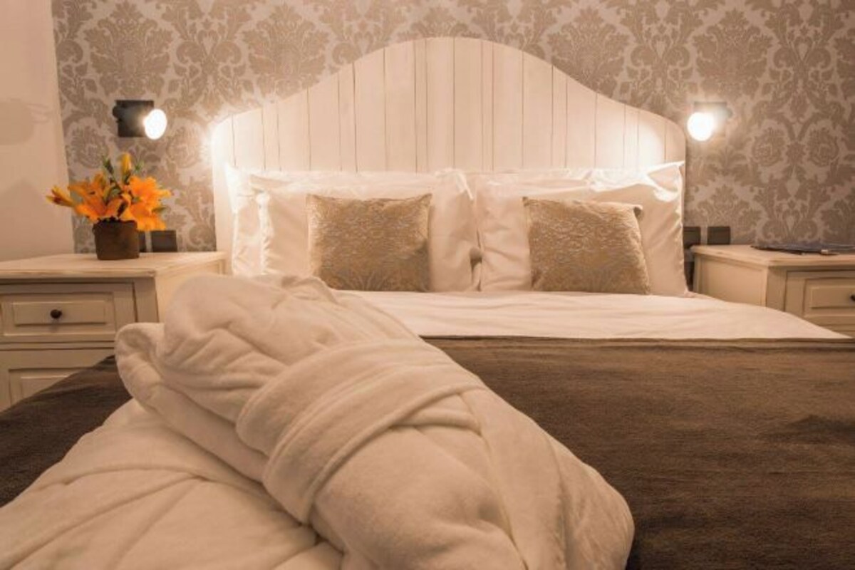 A neatly made bed with white linens and two pillows, a bedside table with a vase of flowers, and a wall with a patterned wallpaper.