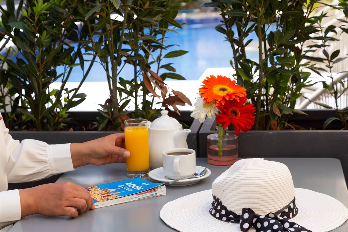 Une table avec un livre, une théière, une tasse de café, un verre de jus d’orange, un chapeau blanc et une fleur.