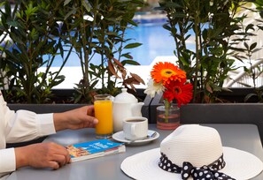 Una mesa con un libro, una tetera, una taza de café, un vaso de jugo de naranja, un sombrero blanco y una flor.