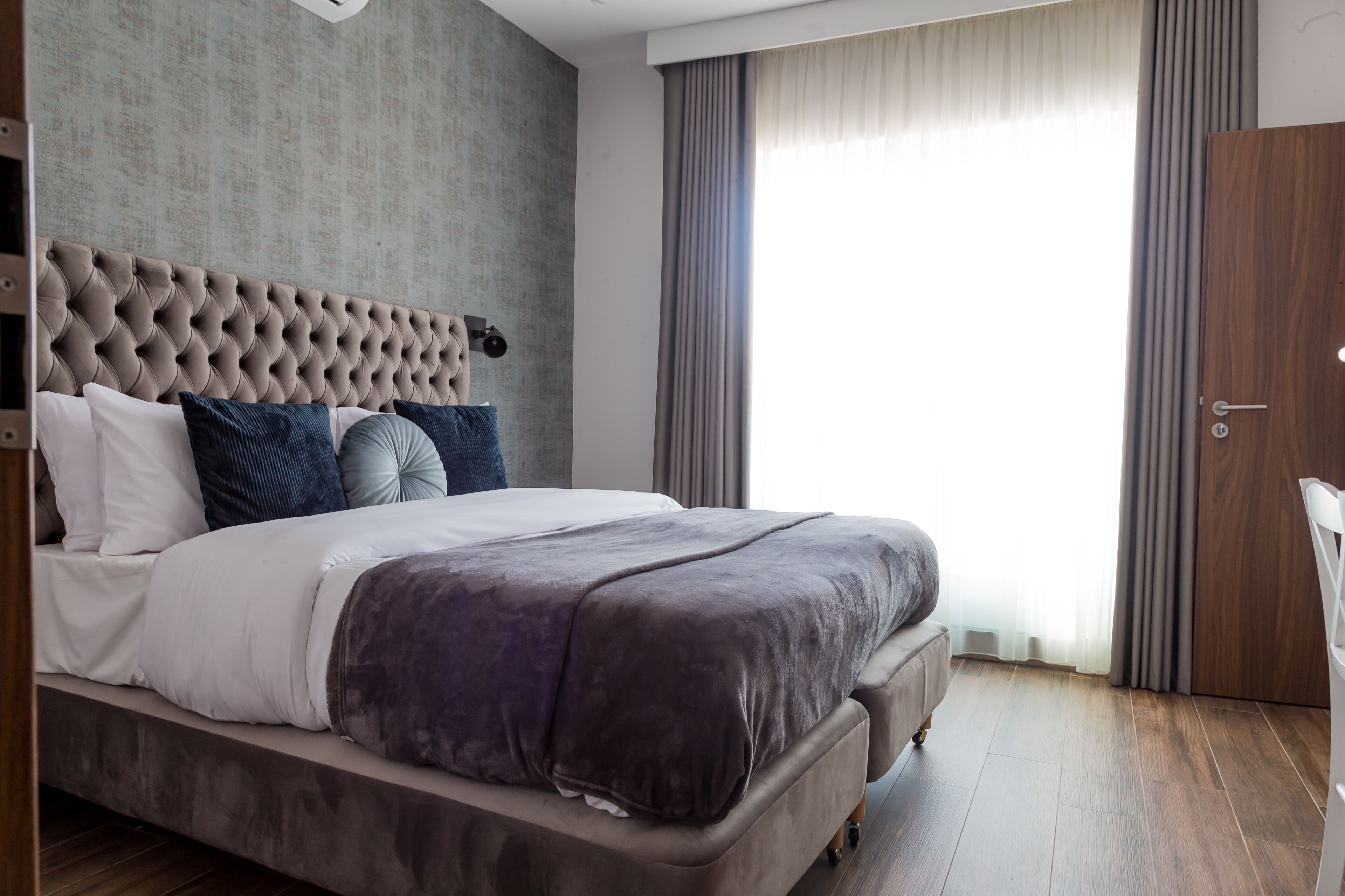 A modern hotel room with a large bed, a tufted headboard, and a view of the outdoors through a large window with curtains.