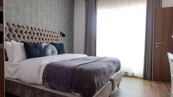 A modern hotel room with a large bed, a tufted headboard, and a view of the outdoors through a large window with curtains.