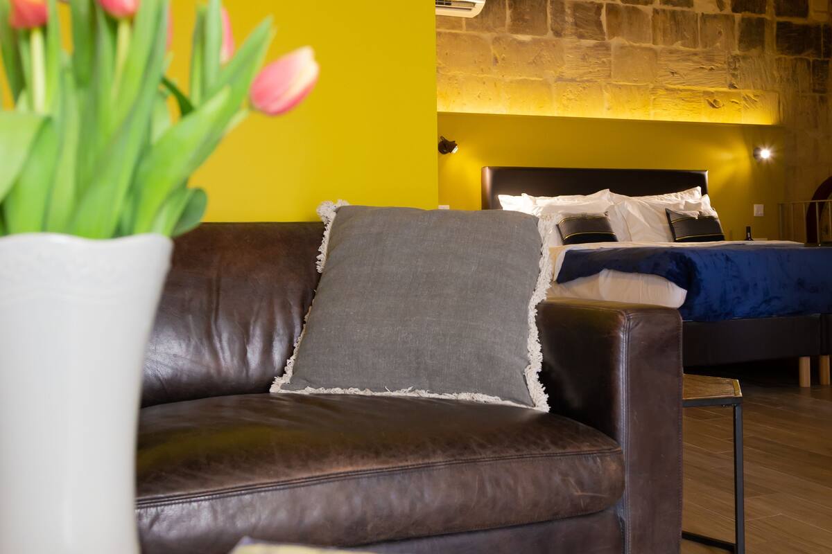 Un salon moderne avec un canapé en cuir marron, un mur d’accent jaune et un lit doté d’une tête de lit bleue.