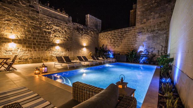 A swimming pool with lounge chairs and a stone wall. - De Rohan B&B (Zebbug)