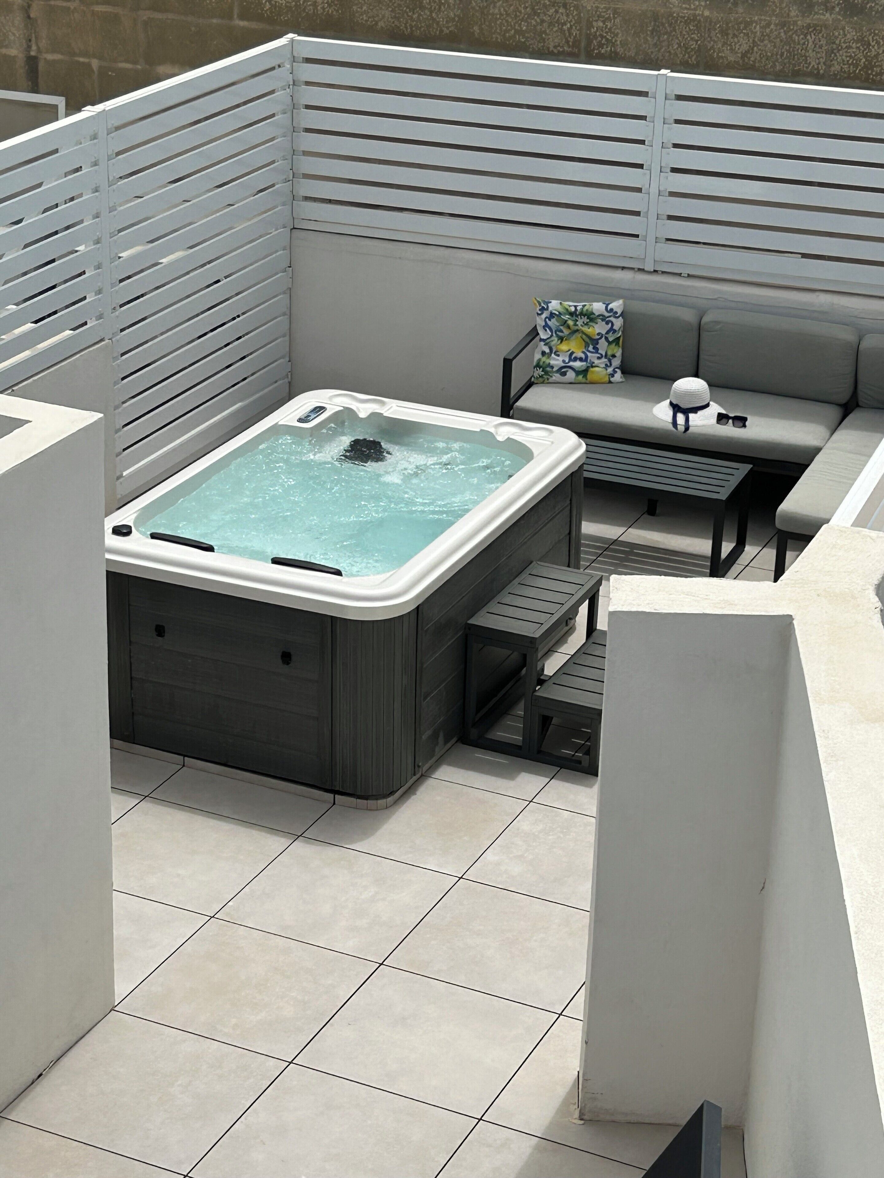 A hot tub in a covered outdoor area with a gray sofa and a small table.