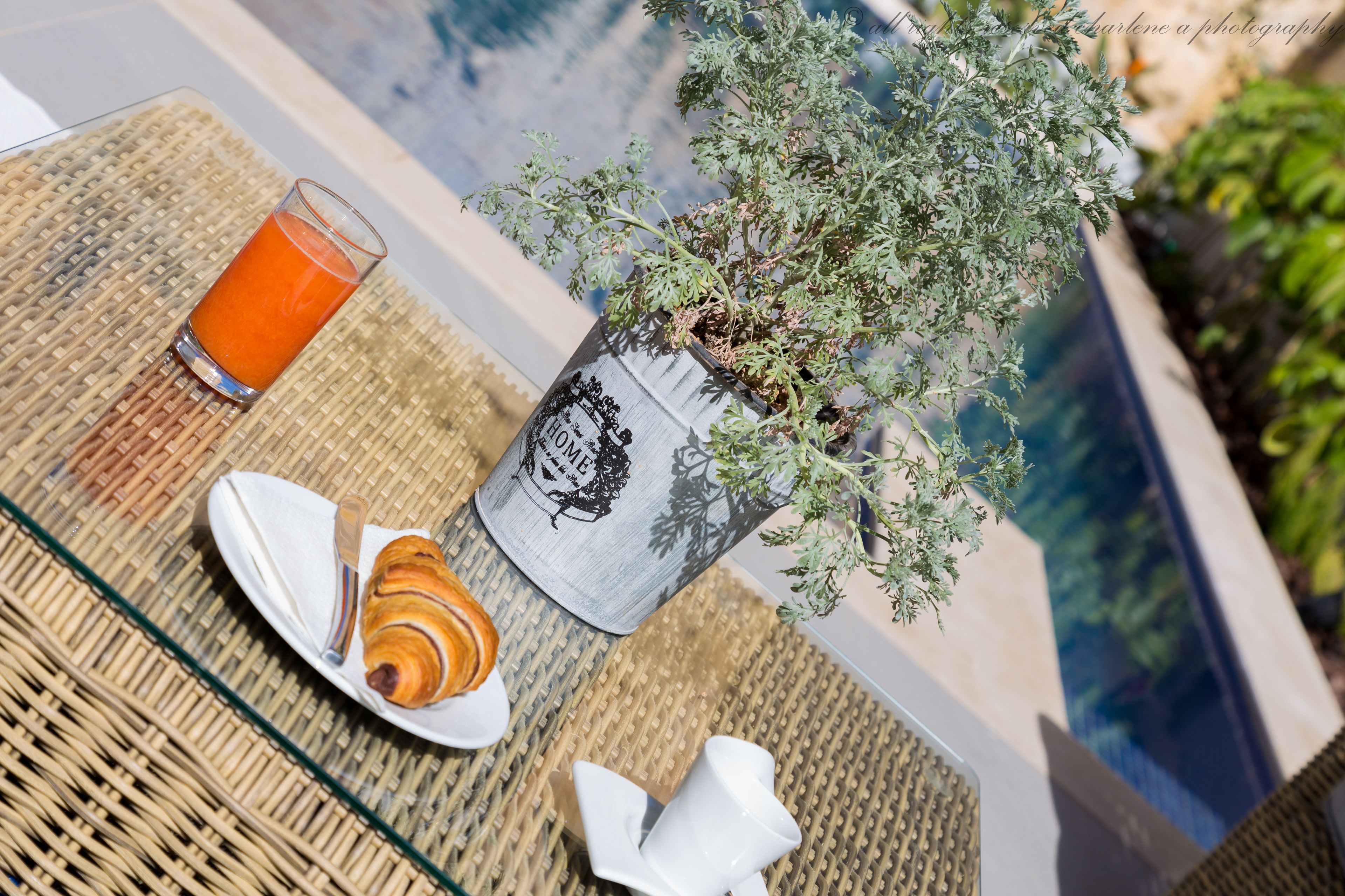Une table mise pour le petit-déjeuner avec un croissant, un verre de jus d’orange et une plante en pot, le tout placé près d’une piscine.