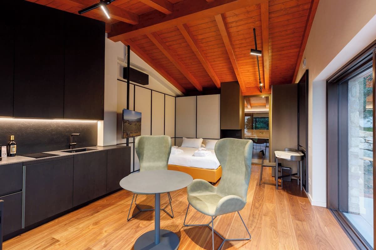 Ein modernes Hotelzimmer mit Holzboden, einem großen Bett, einer Essecke mit Stühlen, einer Küche mit Spüle und einem großen Fenster.