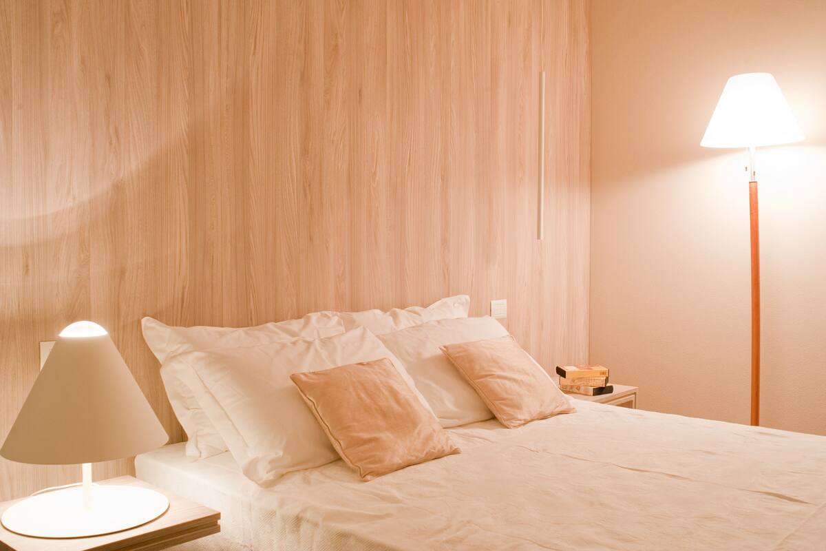 Ein ordentlich bezogenes Bett mit weißen und beigen Kissen, einer Nachttischlampe und einem Nachttisch mit einer Schachtel.