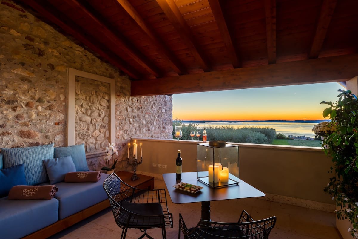 Eine Steinsäule, eine Holzbalkendecke, ein Balkon mit einem Tisch, eine Couch und Blick auf den Sonnenuntergang.