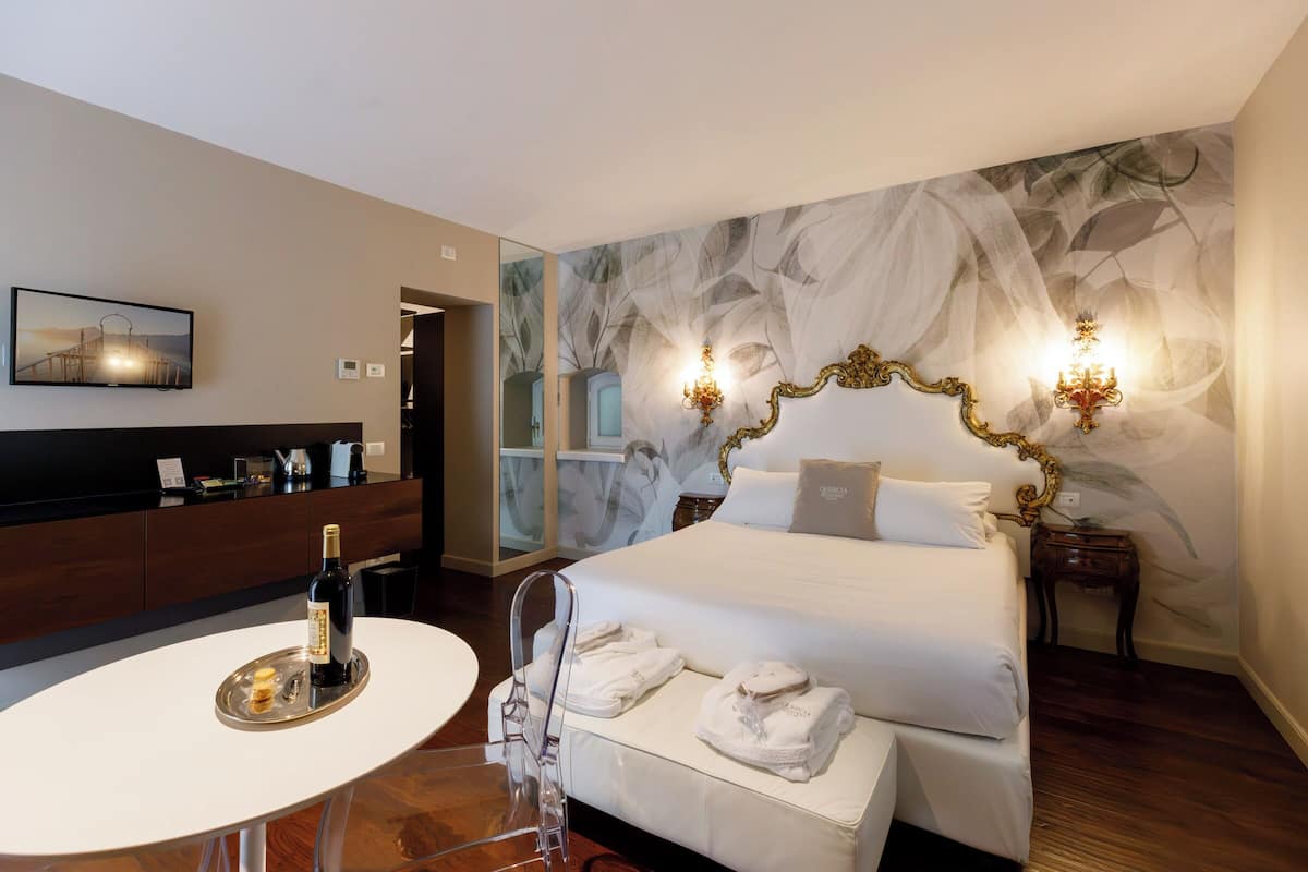 Ein Hotelzimmer mit einem Bett, einem Nachttisch mit Flasche und Gläsern, einem Sessel, einem an der Wand montierten Fernseher und einer Wanddekoration.