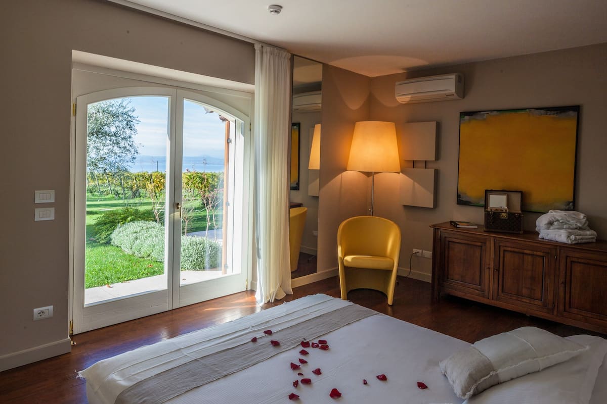 Ein Hotelzimmer mit Bett, Sessel, Lampe, Schminktisch und Blick auf einen Garten durch eine Schiebetür.