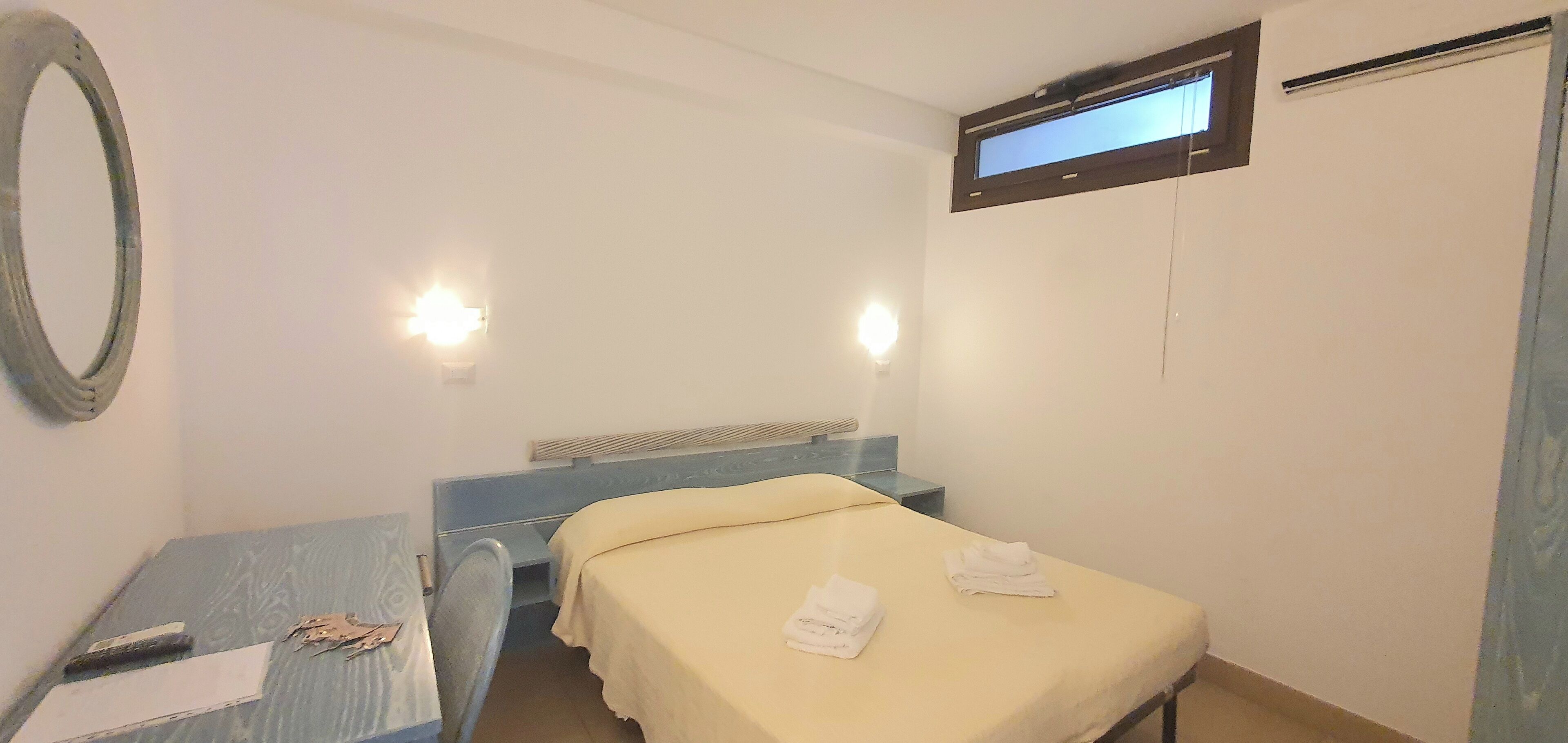 Ein schlichter, kleiner Raum mit einem Bett, einem Schreibtisch und einem Spiegel. Zwei Wandleuchten und ein Fenster mit Jalousie sind vorhanden.