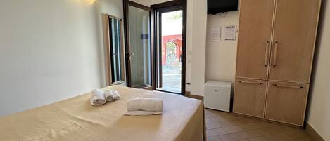 A hotel room with a bed, a wardrobe, a small refrigerator, and a door leading outside.