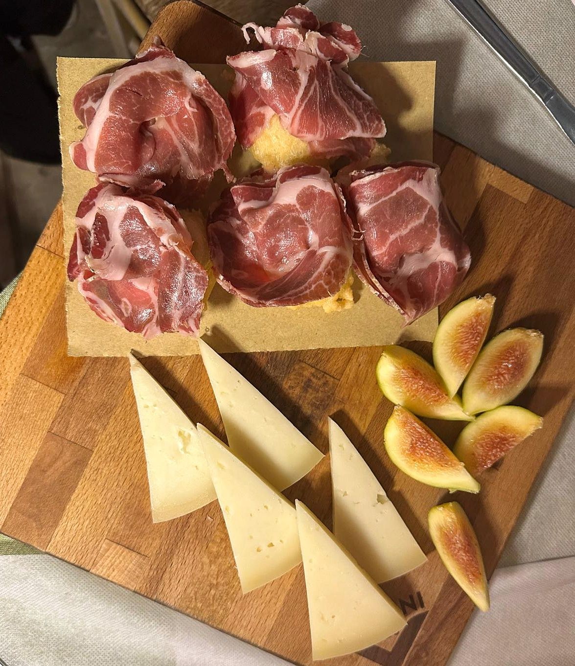 A wooden cutting board with sliced cheese, prosciutto, and figs.
