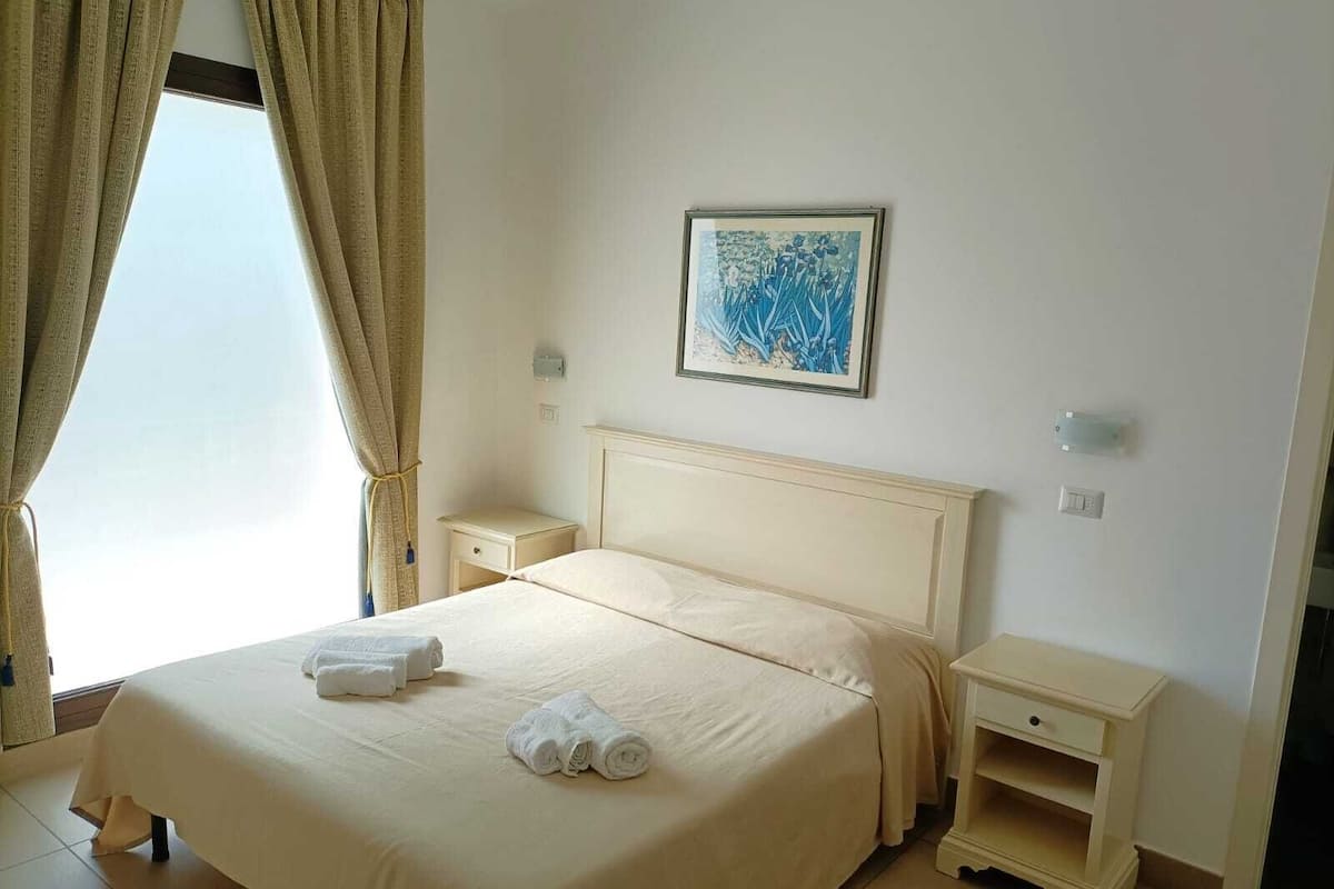 Une chambre d’hôtel avec un lit, deux tables de chevet, une commode, une fenêtre avec des rideaux et un tableau encadré au mur.