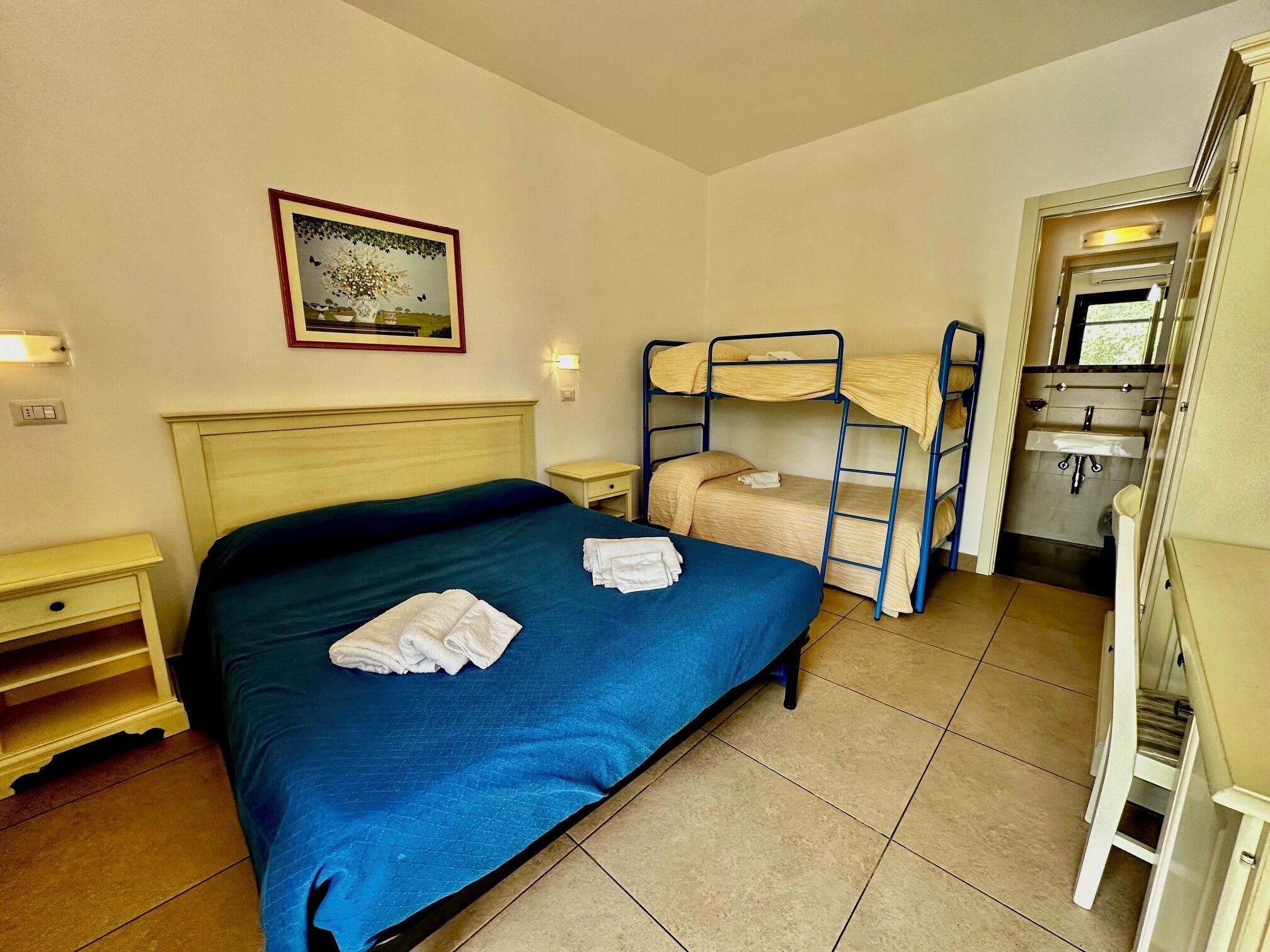 Habitación de hotel con cama individual, literas, una pequeña mesita de noche, lámpara de pared, un cuadro enmarco y baño con lavabo y espejo.