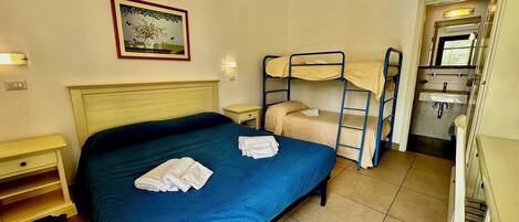 Habitación de hotel con cama individual, literas, una pequeña mesita de noche, lámpara de pared, un cuadro enmarco y baño con lavabo y espejo.