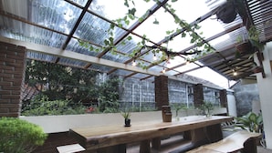 Un patio interior con una mesa y bancos de madera, rodeado de plantas en macetas y un recinto de cristal que ofrece vistas a la vegetación exterior.