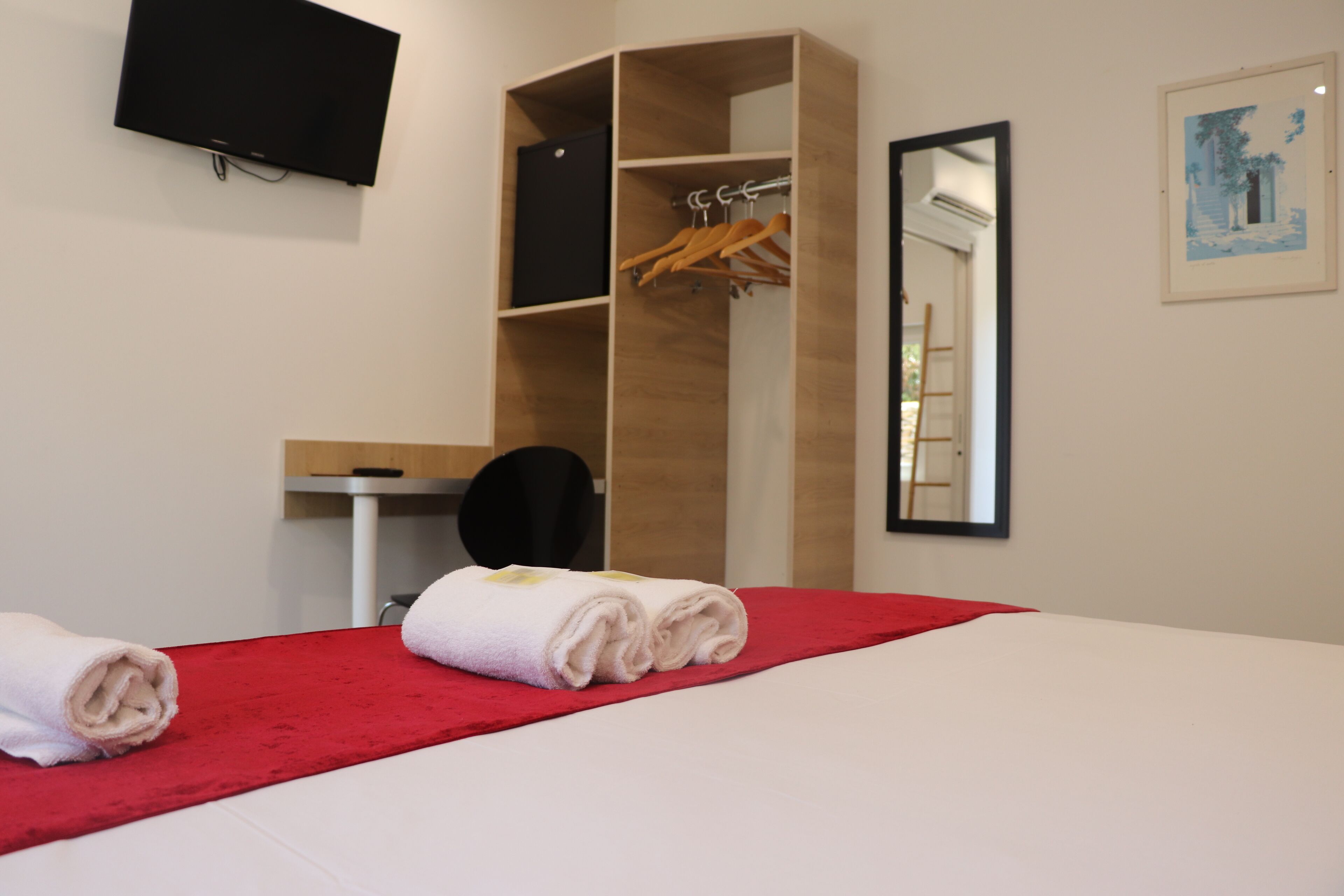 Une chambre d’hôtel avec un lit, une couverture rouge, une armoire en bois, un bureau avec une chaise, un téléviseur à écran plat et un tableau encadré accroché au mur.