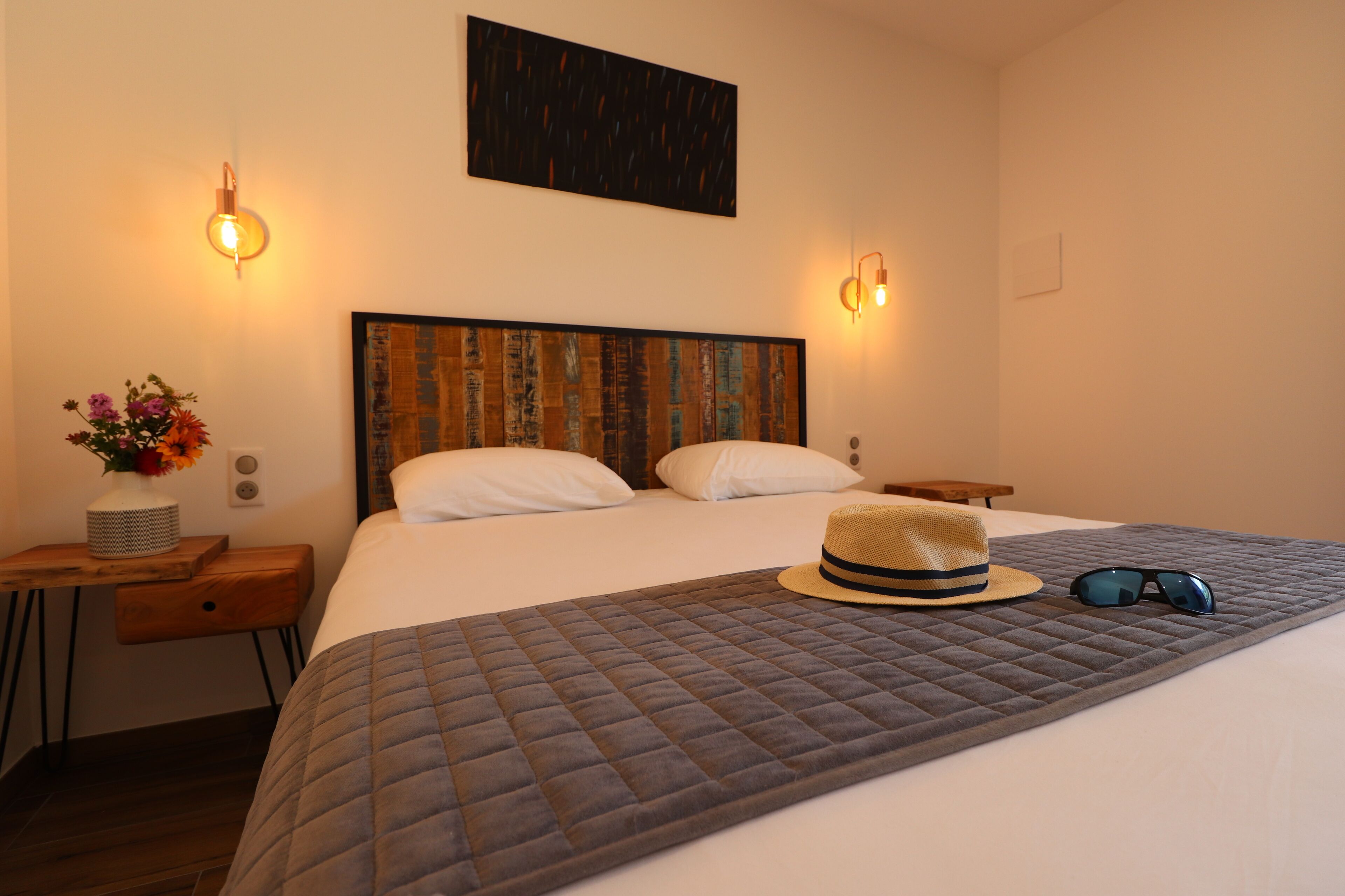 A neatly made bed with a quilted blanket, a wooden bedside table with a vase of flowers, and a wall-mounted light fixture.
