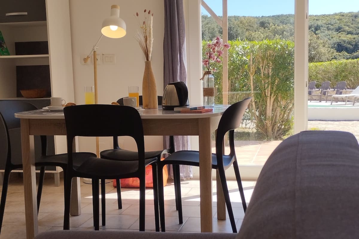 A modern living room with a sofa, a dining table with chairs, a lamp, and a view of a garden through a large window.