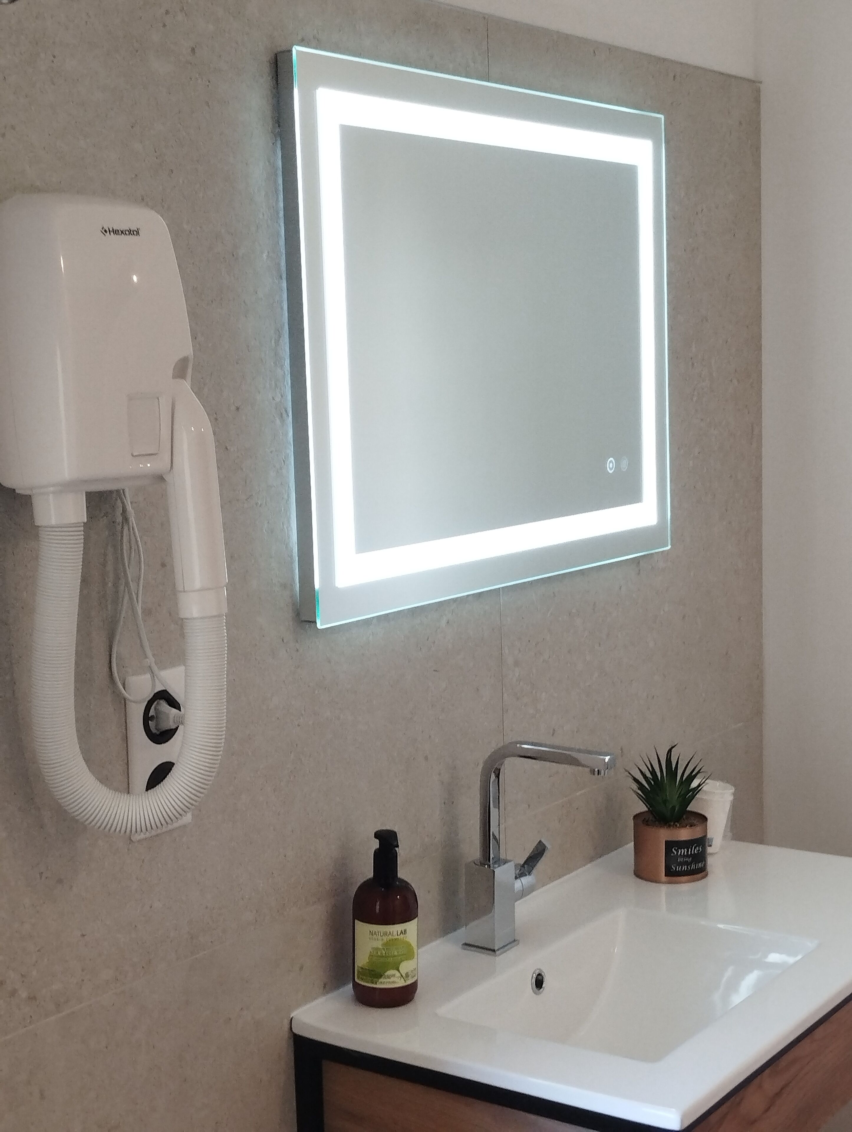 Une salle de bain avec un lavabo blanc, un miroir doté d’un éclairage à LED, un sèche-cheveux, un flacon de savon naturel de laboratoire et une petite plante en pot.