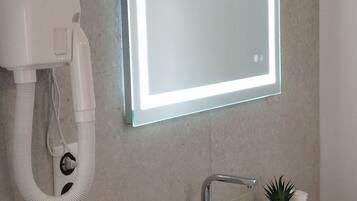 Een badkamer met een wit wastafelmeubel, een spiegel met LED-verlichting, een haardroger, een fles natuurlijke laboratoriumzeep en een kleine potplant.
