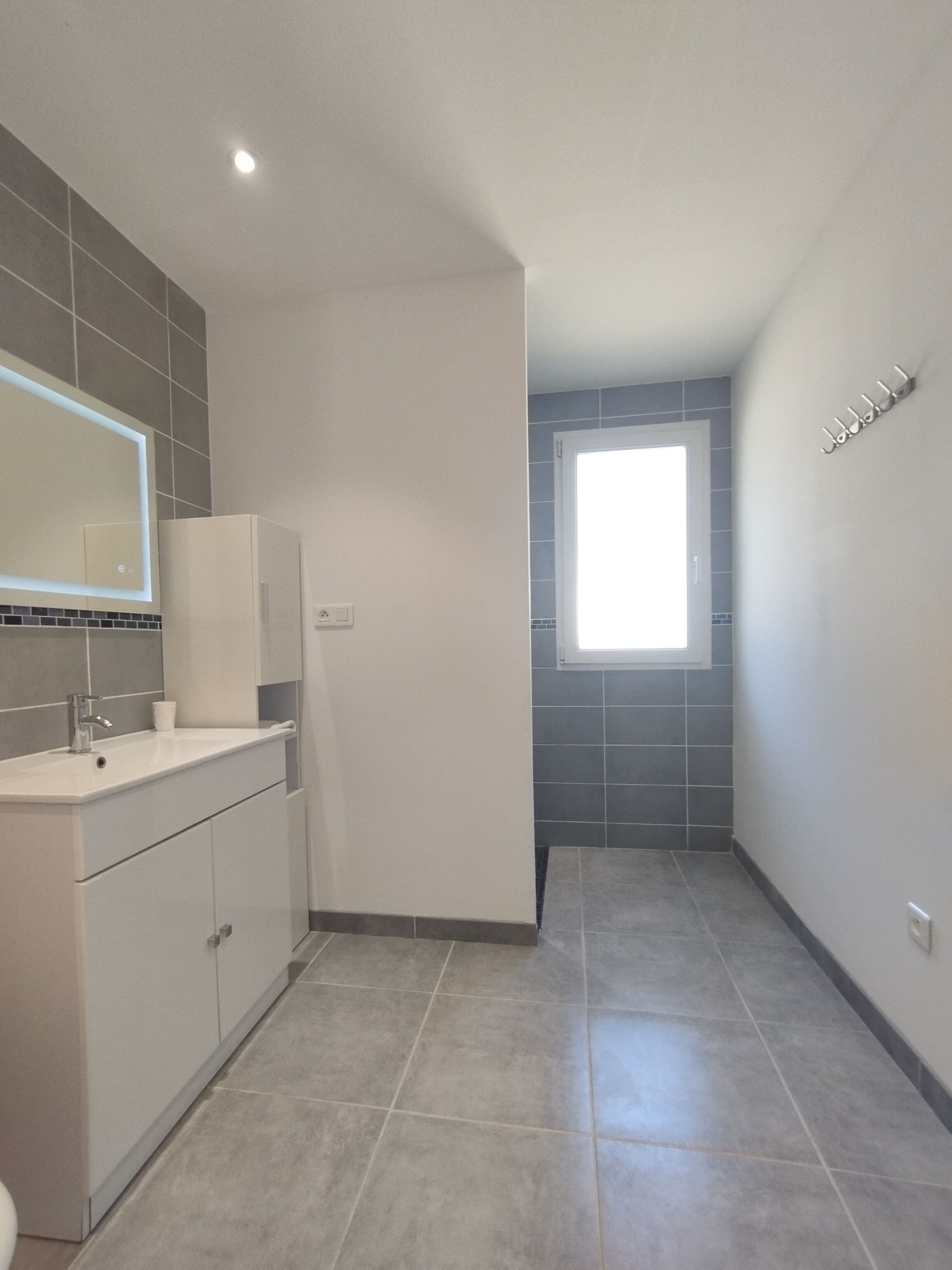 A modern bathroom with a white sink, mirror, and cabinets, a small window, and grey tiled flooring.