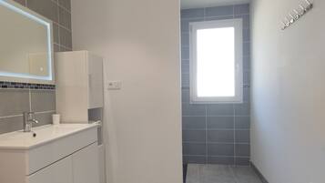 Une salle de bain moderne avec un lavabo blanc, un miroir et des meubles de rangement, une petite fenêtre et un sol carrelé de couleur grise.