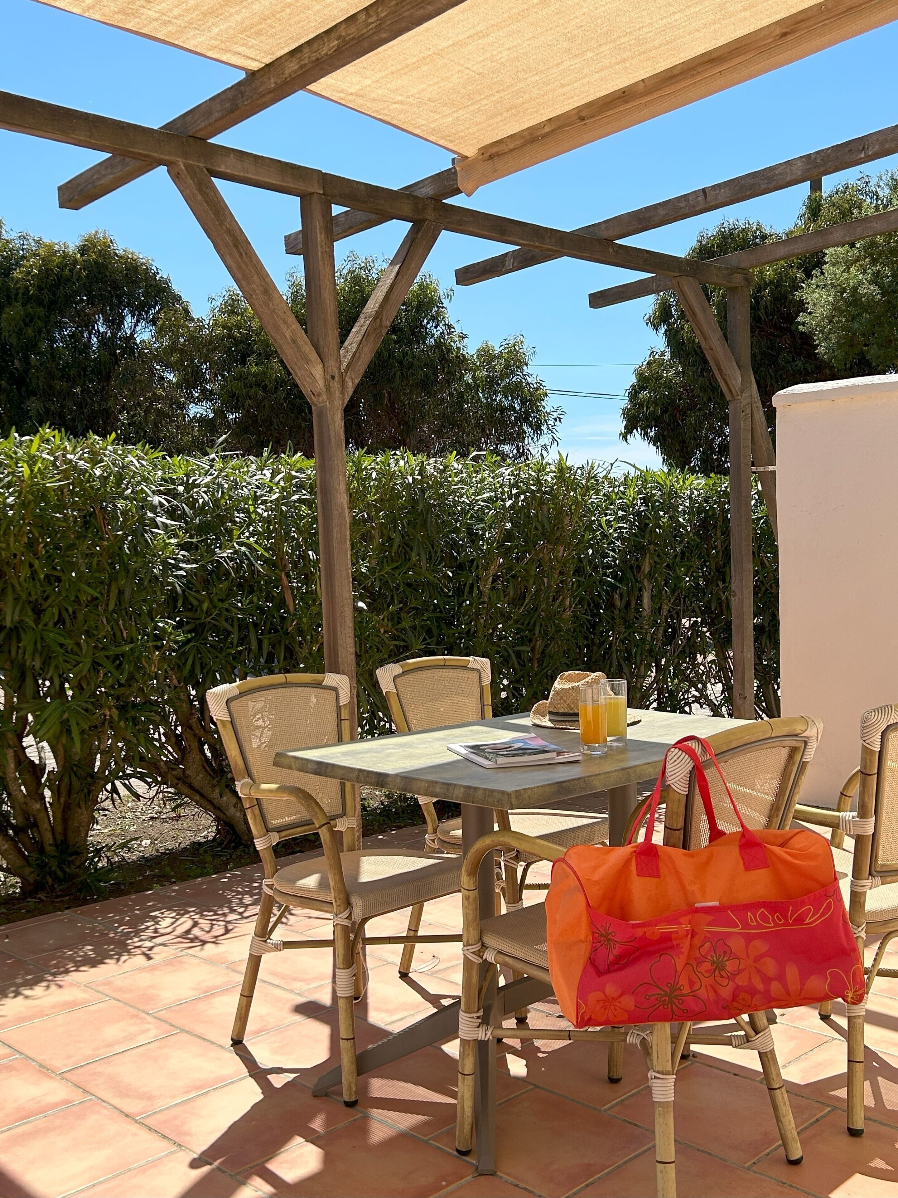 An outdoor patio with a table and chairs, a wooden pergola, and lush greenery in the background.