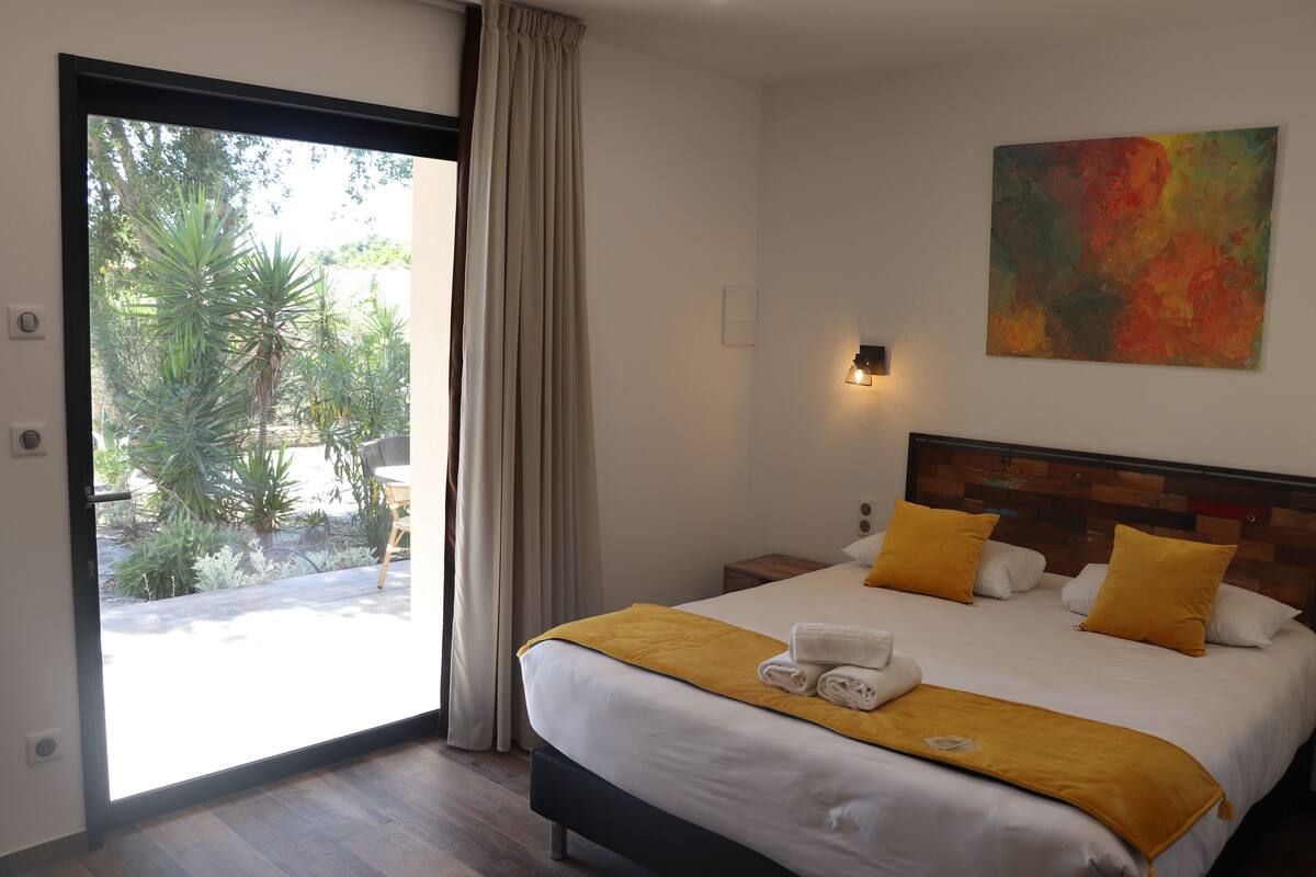 A hotel room with a bed, a large window, a painting, and a view of greenery outside.