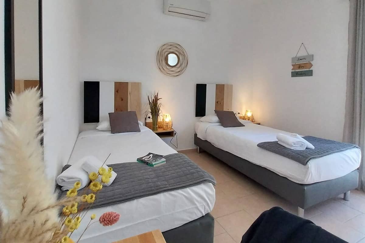A hotel room with two beds, a desk, a mirror, an air conditioner, a hanging light fixture, and a small wall decoration.
