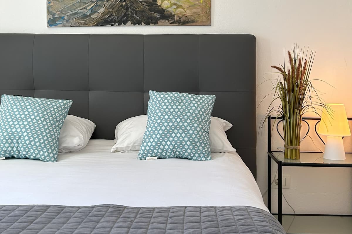A bedroom with a bed, pillows, a headboard, a bedside table with a lamp, and a wall-mounted painting.