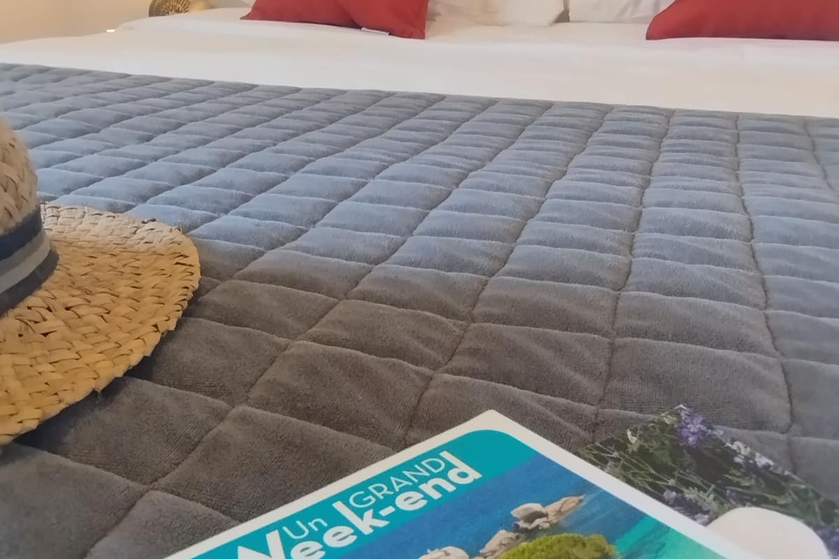 A bed with a grey quilt, red and white pillows, and a straw hat on the side. A book titled "Un Weekend Corse" is visible.
