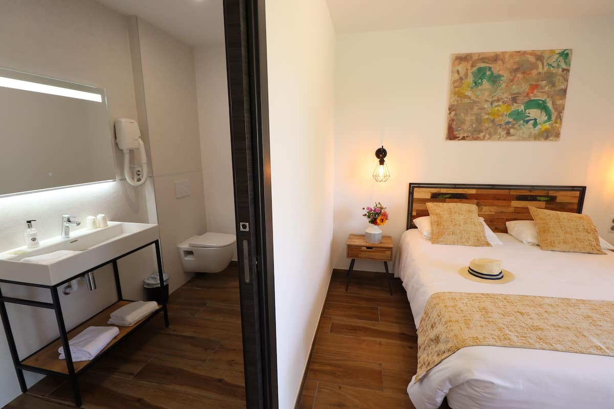 A hotel room with a wooden floor, a white sink with a mirror, a bedside table with a lamp, a bed with pillows, and a painting on the wall.