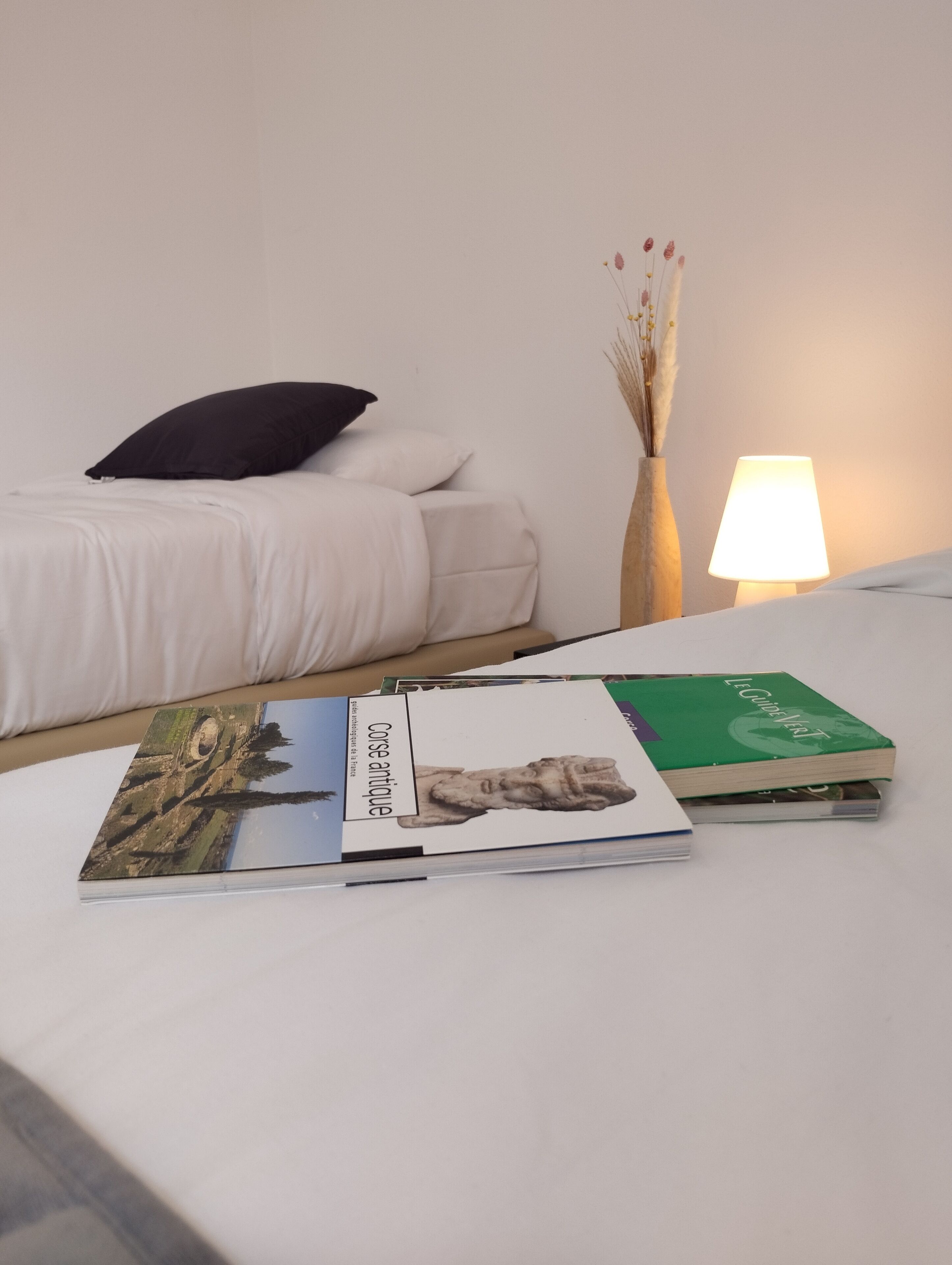 A neatly made bed with white bedding, a black pillow, and a bedside table with a lamp and a vase with dried flowers.