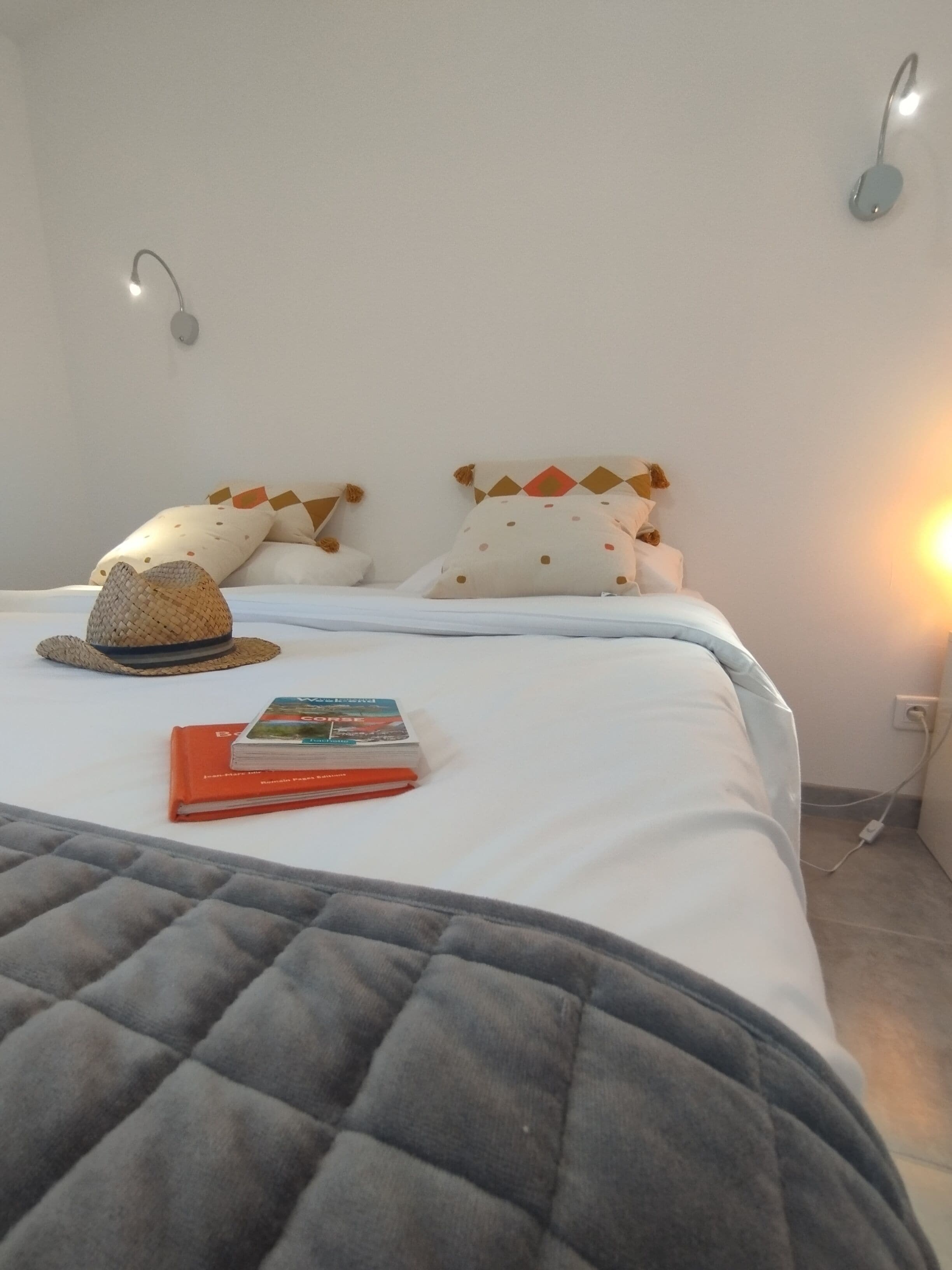 Una cama bien hecha con un edredón gris, un sombrero de paja y un libro de viajes sobre las almohadas.